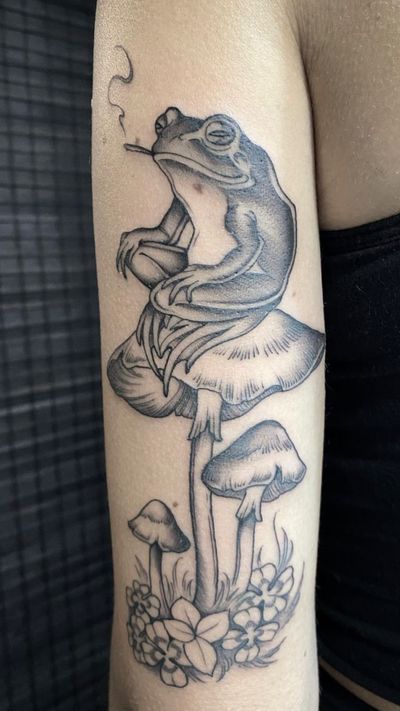 This black and gray tattoo by Amandine Canata features a detailed frog resting on a mushroom, creating a whimsical and nature-inspired design.