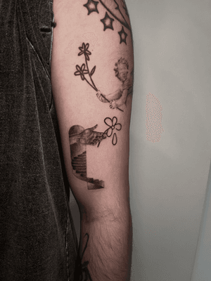 This abstract black and gray tattoo by Lauren features a delicate flower blooming from a hand reaching up a stairway.