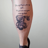 The tattoo depicts a shaded, grayscale teddy bear with a purple bow tie. It's a realistic rendering with soft shading, giving it a sentimental and comforting feel. A purple butterfly hovers nearby, and the text suggests a memorial or remembrance tattoo.
