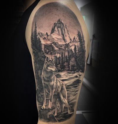 Realistic black and gray tattoo depicting a majestic mountain with a lone wolf overlooking a flowing river and lush forest, by Cara Hayward.