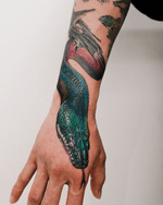 A hyper-realistic White-lipped Python tattoo wraps around the wrist and hand, featuring iridescent scales in deep greens, blues, and purples. The incredible detail and vibrant color gradient give the piece a lifelike, multidimensional appearance.
