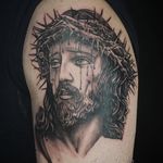 Get a stunning black and gray upper arm tattoo of Jesus wearing a crown of thorns, created by artist Alex Santo.