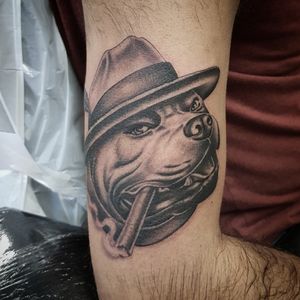 Capture the essence of loyalty and style with this black and gray tattoo of a dog wearing a hat and smoking a cigar on your upper arm. By tattoo artist Dani Mawby.