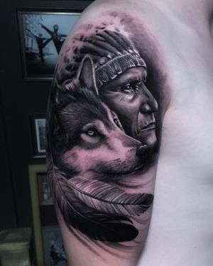 Stunning black and gray upper arm tattoo featuring a realistic illustration of a native man with a wolf and feather motif. By Marcel Oliveira.