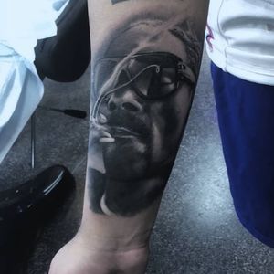Get a detailed black and gray illustration of Snoop Dogg with glasses and cigarette on your forearm by Marcel Oliveira.