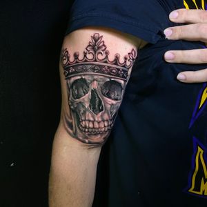 Illustrative black and gray realism tattoo of a skull wearing a crown on upper arm by Marcel Oliveira.