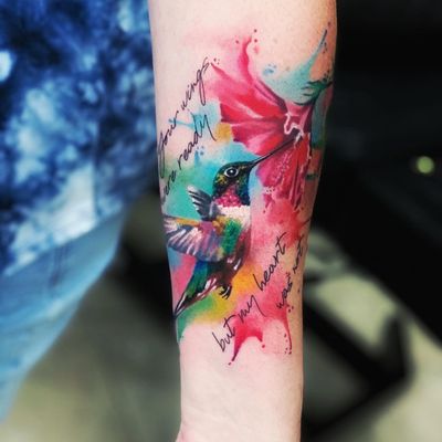 Vibrant watercolor style forearm tattoo featuring a detailed hummingbird and delicate flower, created by the talented artist Marcel Oliveira.