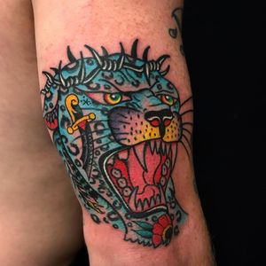 This illustrative tattoo by Felipe Reinoso features a fierce panther surrounded by flowers, a dagger, thorns, and other tattoos on the upper arm.