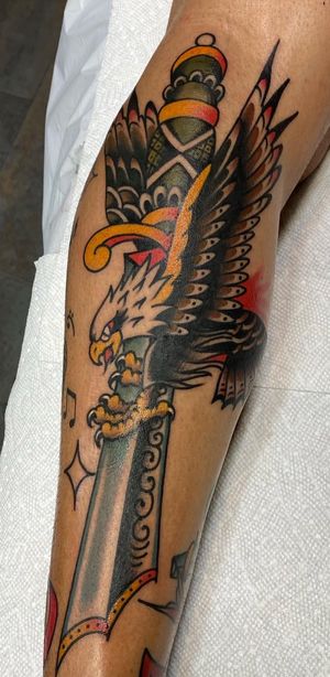 Get inked with a stunning traditional tattoo of an eagle and dagger on your lower leg, skillfully done by Felipe Reinoso.