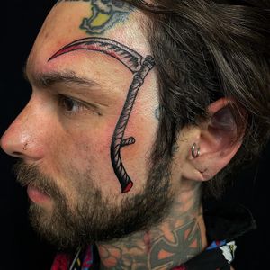 Black and gray tattoo of a scythe on the side of the face, done by Felipe Reinoso. Classic design with a modern twist.