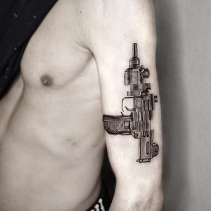 Get a stunning black and gray machine gun tattoo on your upper arm by the talented artist Oek. This detailed design will surely make a bold statement.