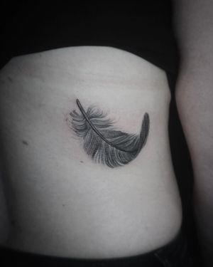 Elegant black and gray fine line feather tattoo delicately placed along the ribs, expertly done by Aygul.