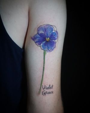 Elegant fine line and watercolor tattoo of a violet flower by Aygul, perfect for a beautiful and feminine upper arm design.