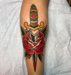 A striking traditional tattoo featuring a vibrant flower and a bold dagger design, expertly crafted by talented artist Daniel Werder on the forearm.