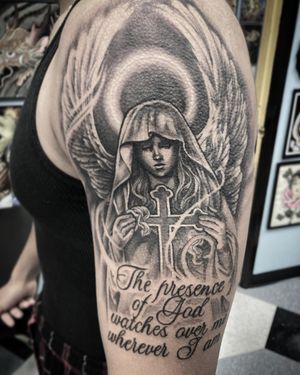 An intricate black and gray tattoo on the upper arm by Rico Dionichi, featuring a cross, angel, and a meaningful quote in illustrative lettering.