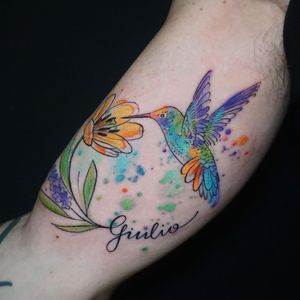 Beautiful watercolor tattoo on upper arm by Aygul, featuring a delicate hummingbird and flower design.