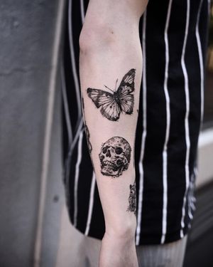 A stunning blackwork and illustrative tattoo featuring a combination of a beautiful butterfly and a sinister skull on the forearm, created by the talented artist Jamie B.