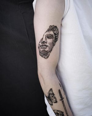 Get a bold and detailed blackwork statue tattoo on your upper arm by the talented artist Jamie B. Embrace the intricate and illustrative style with this unique design.