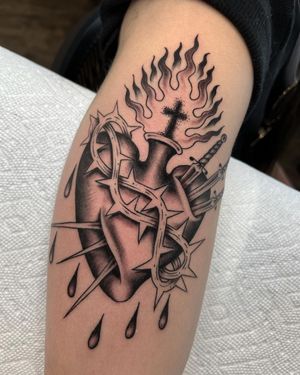 Traditional black and gray tattoo featuring a sacred heart, sword, and cross on the upper arm. By artist Andre Bertoncin.