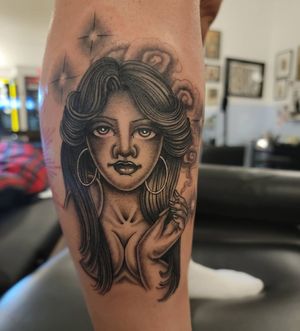 A striking blackwork tattoo of a woman wearing earrings and smoking a cigarette, expertly done on the lower leg by the talented artist Arlene Salinas.