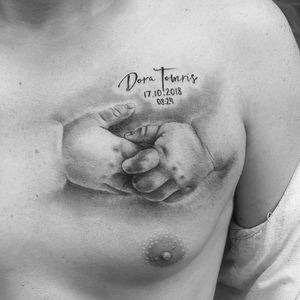 A delicate black and gray illustrative tattoo on the chest featuring a hand, name, and year in small lettering by the talented artist Murat Yılmaz.