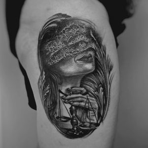 A blackwork tattoo on the upper leg featuring a woman symbolizing justice, blindfolded and holding the scales of balance. By Murat Yılmaz.