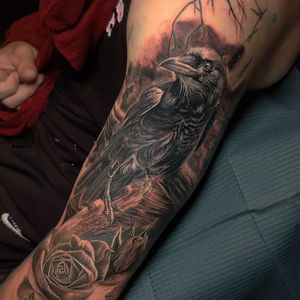 This upper arm tattoo combines blackwork, realism, and illustrative styles featuring a stunning raven perched on a tree with flowers. Located in Chicago, US.