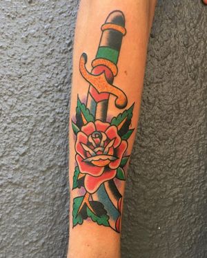 Get inked by Phil Botha with a bold traditional design featuring a flower and dagger motif on your forearm.