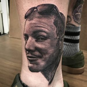 Get a stunning black and gray tattoo of a man with glasses on your lower leg by Sean Ross Fawkes. Perfect mix of realism and lettering.