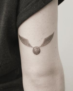 Illustrative black and gray tattoo of golden snitch with wings on upper arm by Martyna Śliwka.
