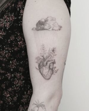 An anatomical heart intertwined with a delicate flower and cloud design, beautifully rendered in black and gray by artist Martyna Śliwka on the upper arm