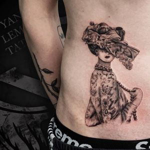 A stunning black and gray illustrative tattoo of a woman with a hat, pearls, necklace, and earrings on the ribs by Yann.