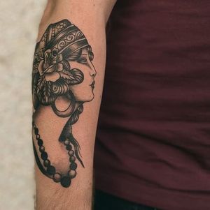 Elegant forearm tattoo by Yura featuring a woman with earrings, bandanna, and necklace surrounded by a beautiful flower motif.