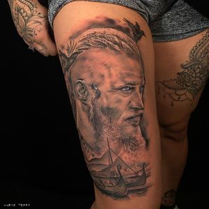 Capture the essence of Travis Fimmel's iconic portrayal of Ragnar with this striking black and gray tattoo on your upper leg. Expertly crafted by Marie Terry.