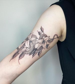 Blackwork tattoo on upper arm featuring a detailed snake intertwined with a blooming flower. By artist Kasia.
