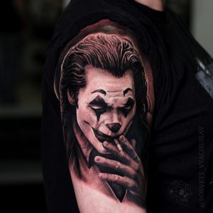 Get a stunning blackwork and realistic upper arm tattoo featuring the joker, cigarette, clown, and the iconic Joaquin Phoenix. Book now!