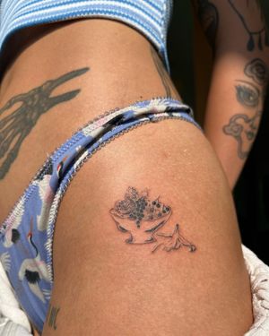 A beautiful blackwork tattoo by Sofi, featuring an illustrative vase filled with vibrant pomegranates on the upper leg