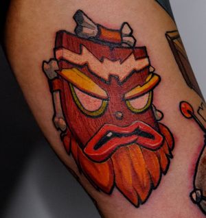 Get a vibrant new school tattoo of the iconic Aku Aku from Crash Bandicoot on your upper leg by the talented artist Denis.