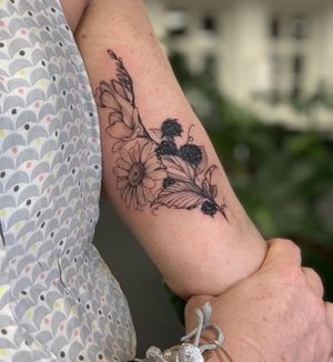 Adorn your upper arm with a striking blackwork and illustrative tattoo of a beautiful combination of flowers and fruits by talented artist Liliia.