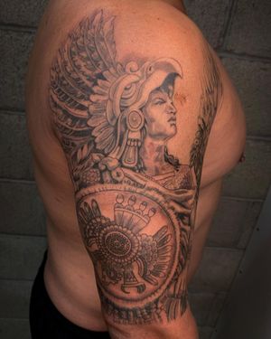 Illustrative upper arm tattoo featuring wings, mask, man, and shield by Angel Chavez.