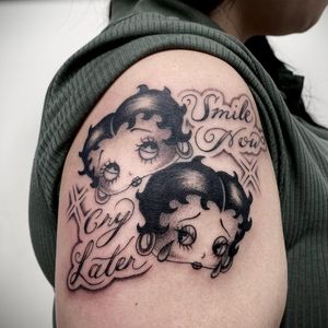 Graceful lettering and detailed portrait of a woman with earrings inspired by Betty Boop, beautifully rendered in black and gray on the upper arm by Sasha Ignarski.