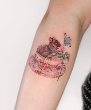 Experience the dynamic artistry of Juliany Braga with this stunning illustrative tattoo featuring a butterfly, flower, rat, and cup on your forearm.