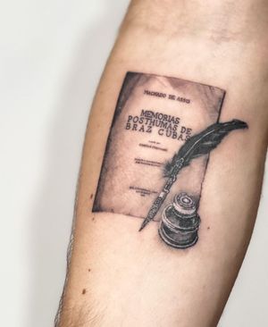 A beautiful black and gray tattoo featuring a feather, book, pen, and ink with a meaningful quote, done by Juliany Braga.