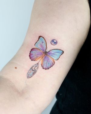 Transform your upper arm with a stunning realistic tattoo of a butterfly and crystal by artist Juliany Braga. The intricate details of this piece will surely make it a standout.