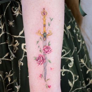 This beautiful forearm tattoo by Juliany Braga features a stunning illustration of a flower intertwined with a sword design.