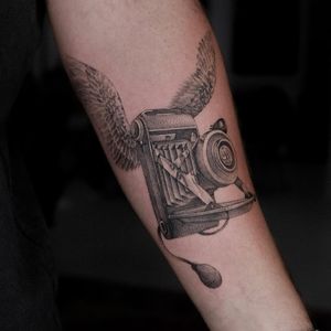 Capture life's moments with this blackwork and realistic tattoo featuring a camera and wings, by artist Oscar Jesus.