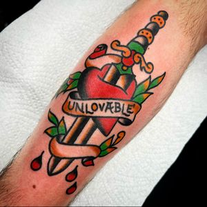Traditional small lettering forearm tattoo by Alessandro Lanzafame featuring a heart and dagger motif.