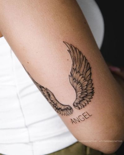 Get inked with a delicate fine line angel wings tattoo by Kateryna Tytarenko. Small lettering adds a personalized touch.