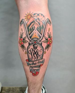 Liza Vettaa's illustrative traditional tattoo on lower leg featuring a sun, mountains, tree, flower, road, quote, and motorcycle.