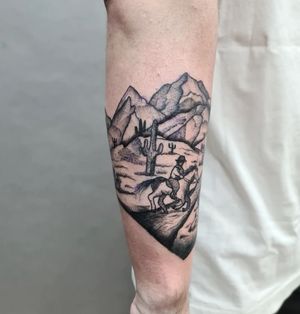 Capture the spirit of the wild west with this forearm tattoo featuring mountains, a horse, cactus, man, and hat in bold blackwork style by Liza Vettaa.